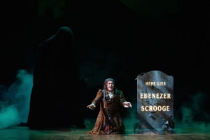 David Studwell. Photo by Jamie Kraus Photography. The Ghost of Christmas Yet to Come looms in the background as Scrooge kneels in despair before a gravestone inscribed with “Here Lies Ebenezer Scrooge.” Wisps of fog surround the stage, creating an eerie atmosphere.