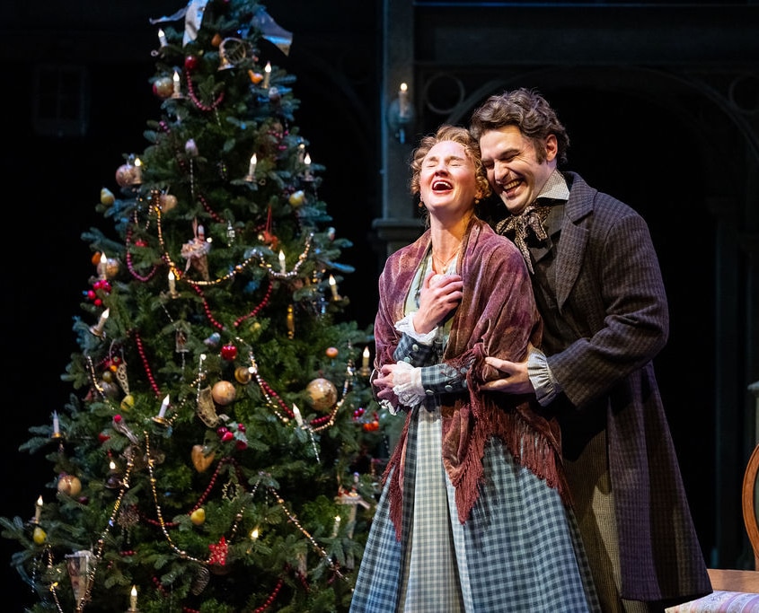 Chloe McLeod and Brian Bohlender. Photo by Jamie Kraus Photography. Belle and her husband stand close together in front of a beautifully decorated Christmas tree with ornaments and garlands. The intimate moment suggests a heartfelt exchange in a warmly lit setting.
