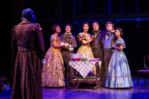 The cast of A Christmas Carol. Photo by Jamie Kraus Photography. A group in elaborate period costumes gathers around a table with a lace cloth, expressing surprise and joy. The scene is warmly lit, conveying a festive atmosphere.