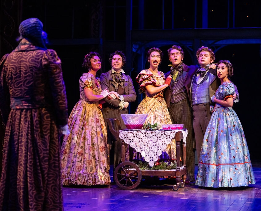 The cast of A Christmas Carol. Photo by Jamie Kraus Photography. A group in elaborate period costumes gathers around a table with a lace cloth, expressing surprise and joy. The scene is warmly lit, conveying a festive atmosphere.