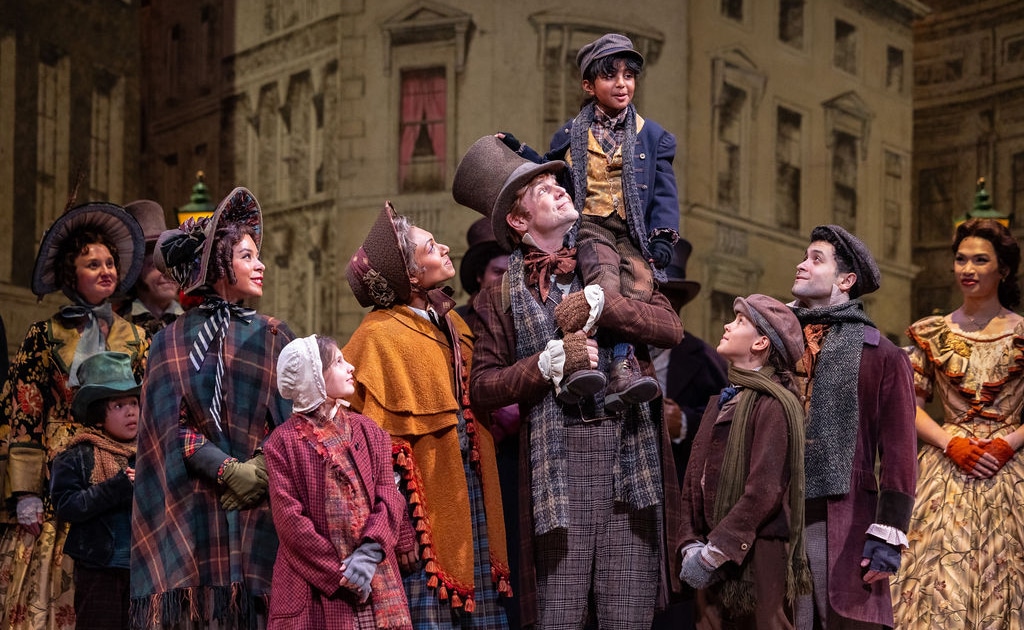 The Cratchit family stands together on stage in Victorian-era clothing. Bob Cratchit stands in the center joyfully holding Tiny Tim of his shoulders.
