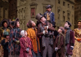The Cratchit family stands together on stage in Victorian-era clothing. Bob Cratchit stands in the center joyfully holding Tiny Tim of his shoulders.