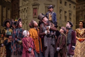 Photo Cred: The cast of A Christmas Carol. Photo by Jamie Kraus Photography. The Cratchit family stands together on stage in Victorian-era clothing. Bob Cratchit stands in the center joyfully holding Tiny Tim of his shoulders.