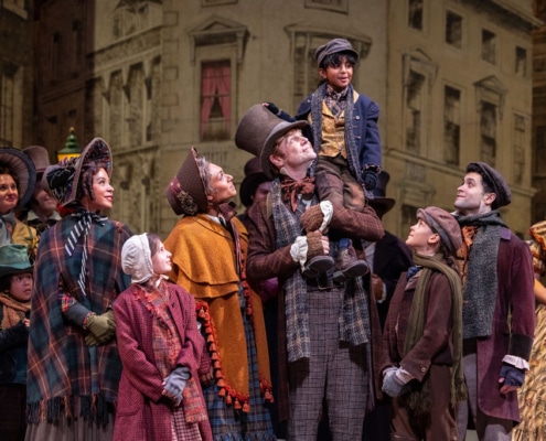 The Cratchit family stands together on stage in Victorian-era clothing. Bob Cratchit stands in the center joyfully holding Tiny Tim of his shoulders.