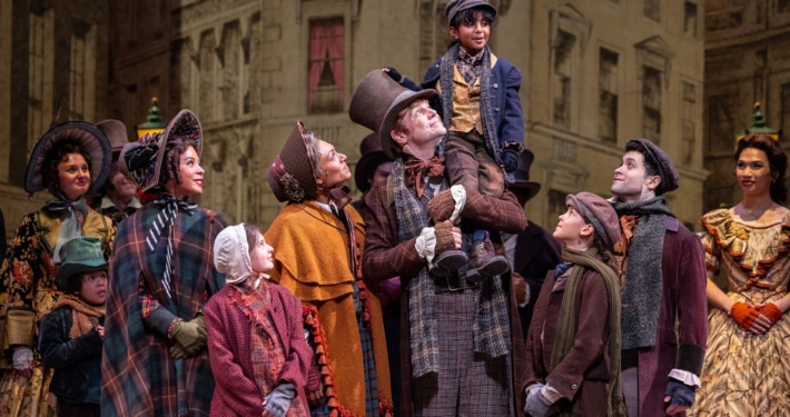 The Cratchit family stands together on stage in Victorian-era clothing. Bob Cratchit stands in the center joyfully holding Tiny Tim of his shoulders.