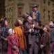 The Cratchit family stands together on stage in Victorian-era clothing. Bob Cratchit stands in the center joyfully holding Tiny Tim of his shoulders.