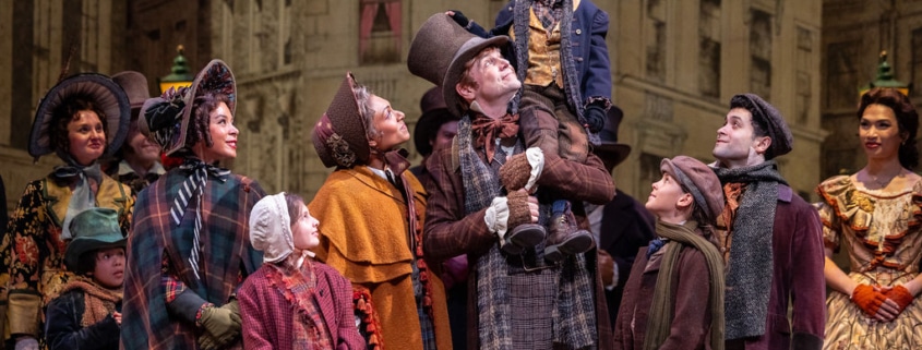 The Cratchit family stands together on stage in Victorian-era clothing. Bob Cratchit stands in the center joyfully holding Tiny Tim of his shoulders.