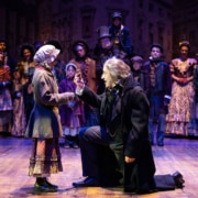 A stage scene depicts a Victorian setting with Scrooge kneeling in front of a girl in period costume. The cast of A Christmas Carol watches on.