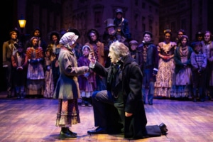 Cierra Banks and David Studwell. Photo by Jamie Kraus Photography. A stage scene depicts a Victorian setting with Scrooge kneeling in front of a girl in period costume. The cast of A Christmas Carol watches on.