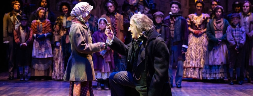 A stage scene depicts a Victorian setting with Scrooge kneeling in front of a girl in period costume. The cast of A Christmas Carol watches on.