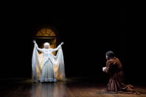 Alia Munsch and David Studwell. Photo by Jamie Kraus Photography. The Ghost of Christmas Past stands in a radiant white gown with her arms raised, illuminated against a dark stage. Scrooge kneels before her in a dress robe kneels.