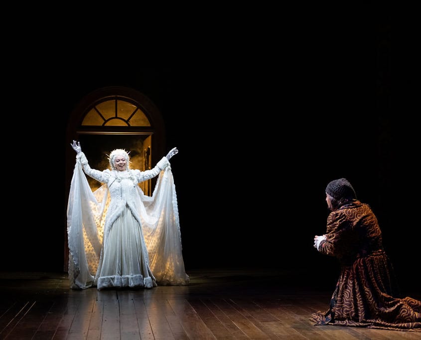 Alia Munsch and David Studwell. Photo by Jamie Kraus Photography. The Ghost of Christmas Past stands in a radiant white gown with her arms raised, illuminated against a dark stage. Scrooge kneels before her in a dress robe kneels.