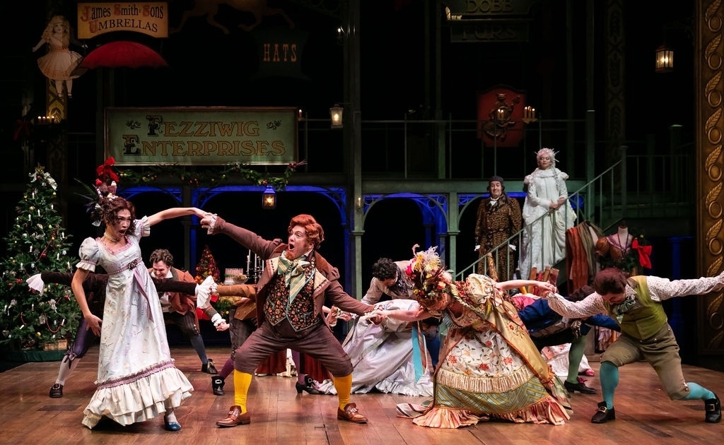 A group of people in colorful Victorian garb dance around the stage as Ebenezar Scrooge and The Ghost of Christmas Past watch