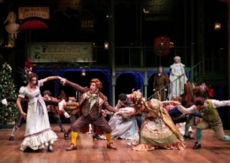 A group of people in colorful Victorian garb dance around the stage as Ebenezar Scrooge and The Ghost of Christmas Past watch