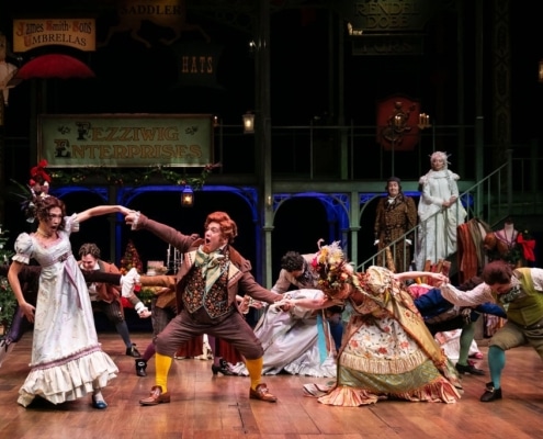 A group of people in colorful Victorian garb dance around the stage as Ebenezar Scrooge and The Ghost of Christmas Past watch