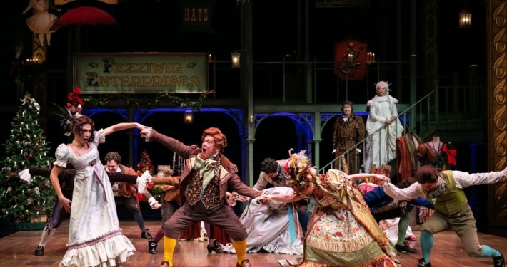 A group of people in colorful Victorian garb dance around the stage as Ebenezar Scrooge and The Ghost of Christmas Past watch