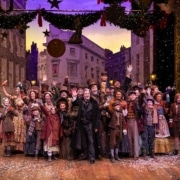 A large group of people wearing Victorian garb stand on stage, smiling and waving at the camera.