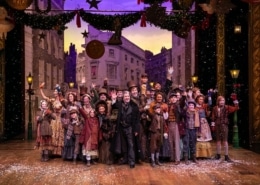 A large group of people wearing Victorian garb stand on stage, smiling and waving at the camera.