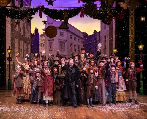A large group of people wearing Victorian garb stand on stage, smiling and waving at the camera.