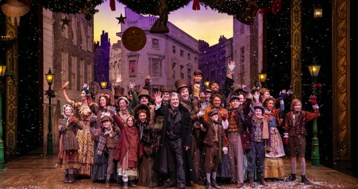 A large group of people wearing Victorian garb stand on stage, smiling and waving at the camera.