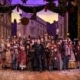 A large group of people wearing Victorian garb stand on stage, smiling and waving at the camera.
