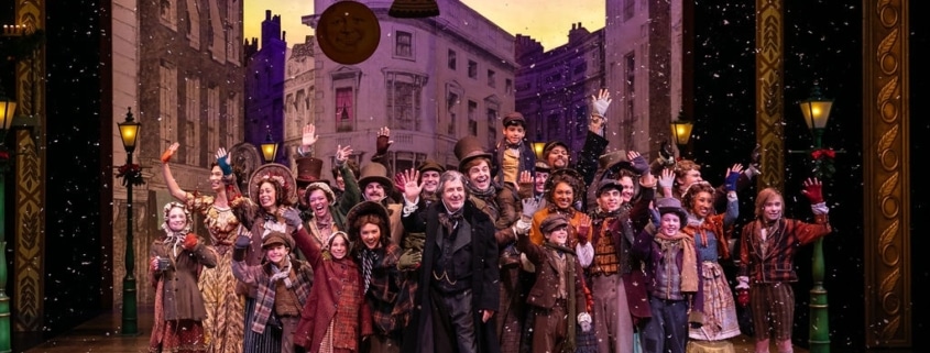 A large group of people wearing Victorian garb stand on stage, smiling and waving at the camera.