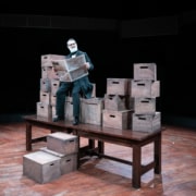 An older gentleman reads a newspaper. He's seated on a stack of wooden boxes, stacked on top of a wooden table.