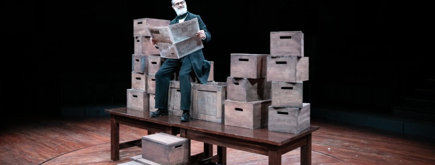 An older gentleman reads a newspaper. He's seated on a stack of wooden boxes, stacked on top of a wooden table.