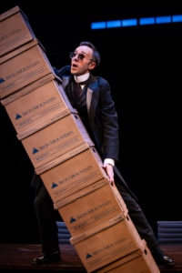 Mayer Lehman stands on a table wearing a three piece suit and sunglasses. He's frantically holding up a leaning, enormous stack of boxes to keep them from tipping.