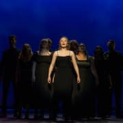 Charlotte stands in the center spotlight. The other nine nominees stand in shadow behind her, forming a pyramid.