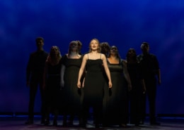 Charlotte stands in the center spotlight. The other nine nominees stand in shadow behind her, forming a pyramid.