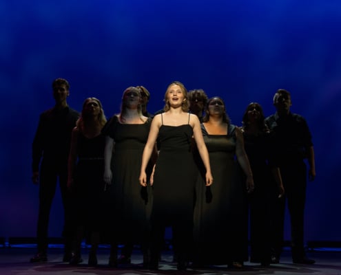 Charlotte stands in the center spotlight. The other nine nominees stand in shadow behind her, forming a pyramid.