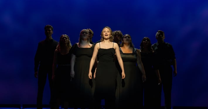 Charlotte stands in the center spotlight. The other nine nominees stand in shadow behind her, forming a pyramid.