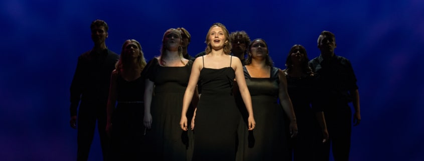 Charlotte stands in the center spotlight. The other nine nominees stand in shadow behind her, forming a pyramid.