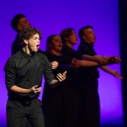 Tate Dewhurst stands in the spotlight, singing with passion. Four other nominees stand behind him, their arms out.