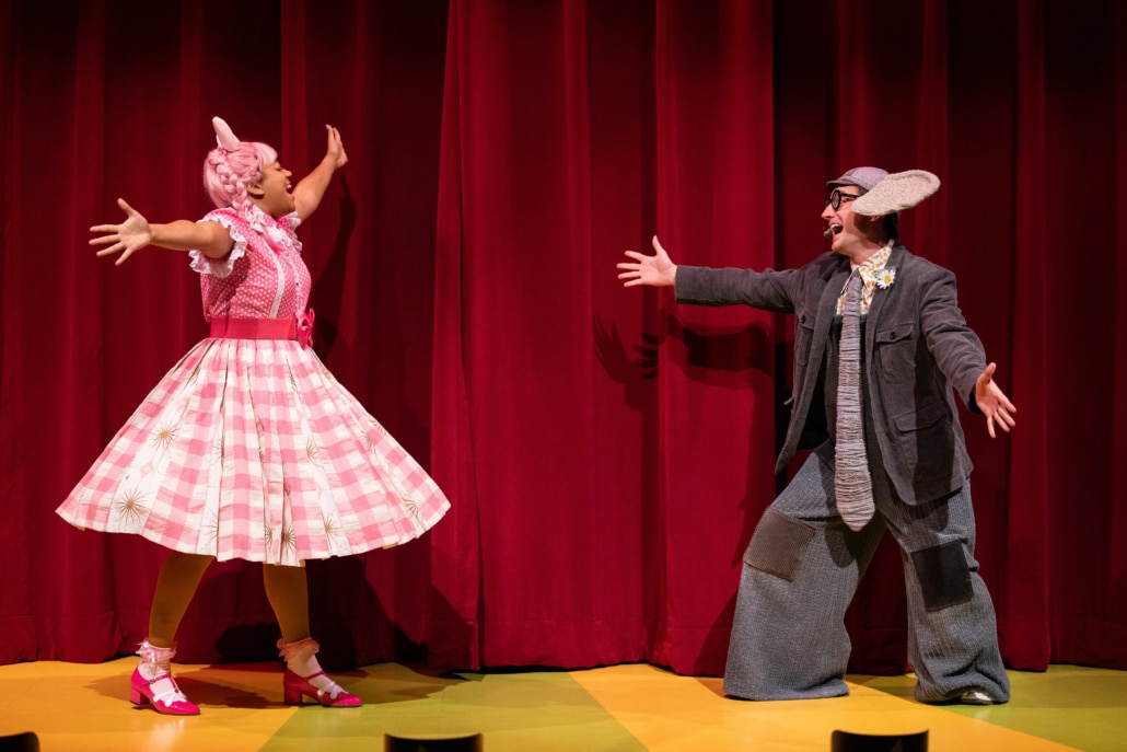 Piggie and Gerald the elephant hold out their arms in greeting, smiling widely. Piggie wears a pink dress and shoes, Gerald is wearing an oversized gray suit.