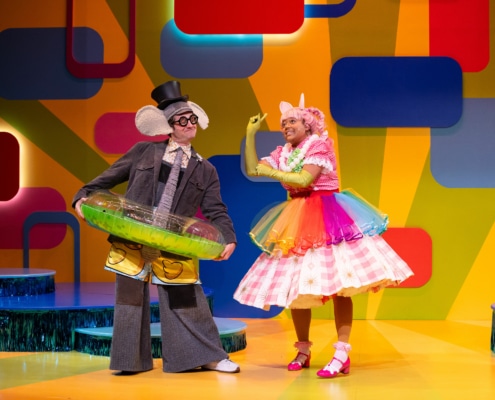 Gerald the elephant and Piggie stand onstage, wearing fun accessories like a pool inner tube, a rainbow tutu, and a top hat.