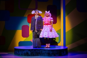 Gerald the Elephant and Piggie hold hands on top of a blue platform singing.