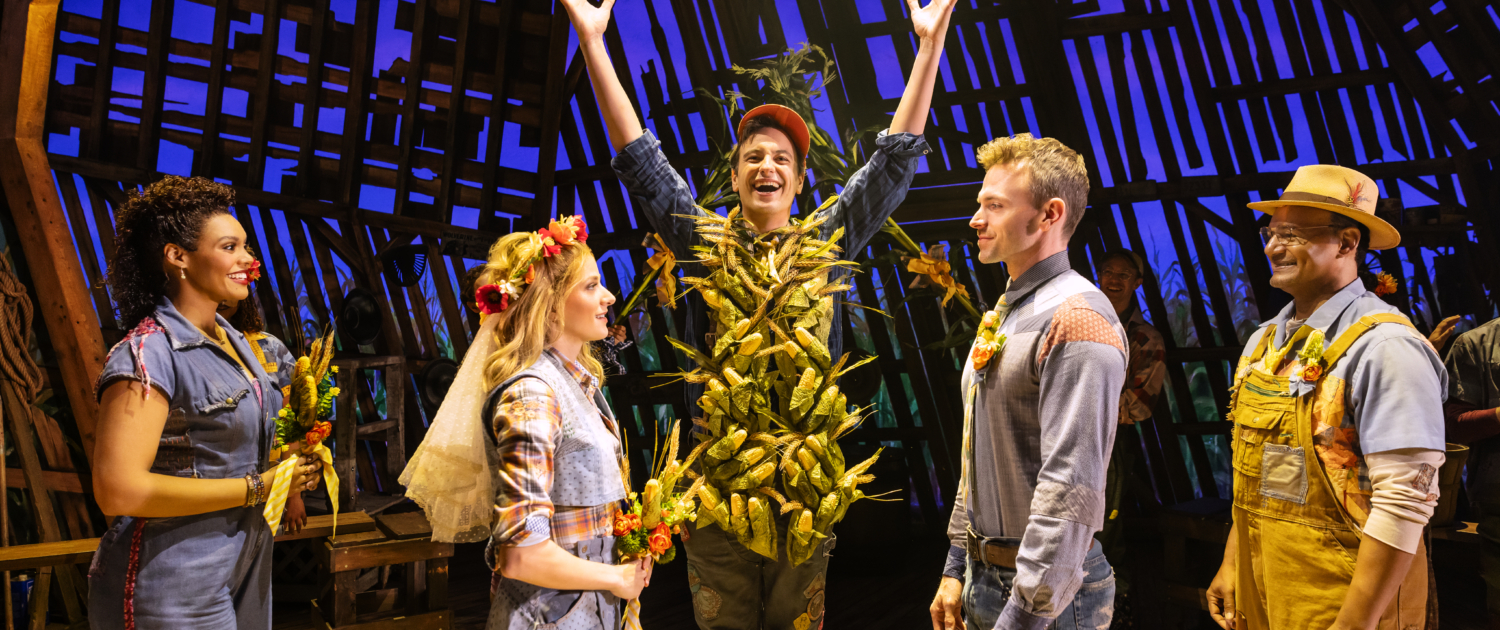 Five characters stand on stage. The three in the center depict a wedding: (l-r) the woman is dressed with a floral headpiece and veil in a blue dress holding flowers and facing a man in jeans and denim shirt, while a central character stands with his hands raised and his torso covered in corn cobs. A woman and man who serve as attendants are on either side.