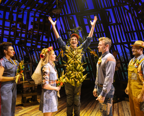 Five characters stand on stage. The three in the center depict a wedding: (l-r) the woman is dressed with a floral headpiece and veil in a blue dress holding flowers and facing a man in jeans and denim shirt, while a central character stands with his hands raised and his torso covered in corn cobs. A woman and man who serve as attendants are on either side.