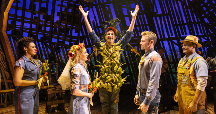 Five characters stand on stage. The three in the center depict a wedding: (l-r) the woman is dressed with a floral headpiece and veil in a blue dress holding flowers and facing a man in jeans and denim shirt, while a central character stands with his hands raised and his torso covered in corn cobs. A woman and man who serve as attendants are on either side.