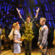 Five characters stand on stage. The three in the center depict a wedding: (l-r) the woman is dressed with a floral headpiece and veil in a blue dress holding flowers and facing a man in jeans and denim shirt, while a central character stands with his hands raised and his torso covered in corn cobs. A woman and man who serve as attendants are on either side.