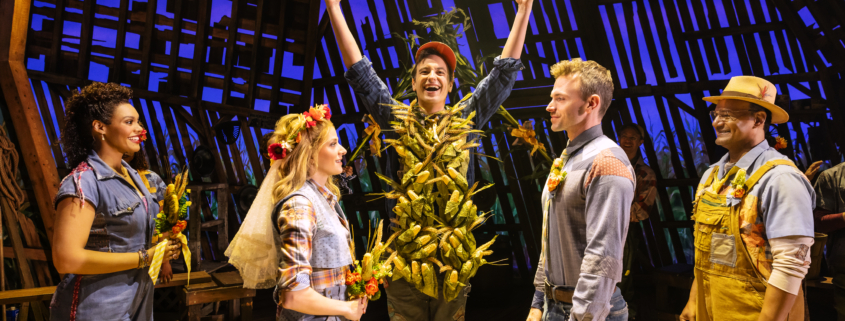 Five characters stand on stage. The three in the center depict a wedding: (l-r) the woman is dressed with a floral headpiece and veil in a blue dress holding flowers and facing a man in jeans and denim shirt, while a central character stands with his hands raised and his torso covered in corn cobs. A woman and man who serve as attendants are on either side.