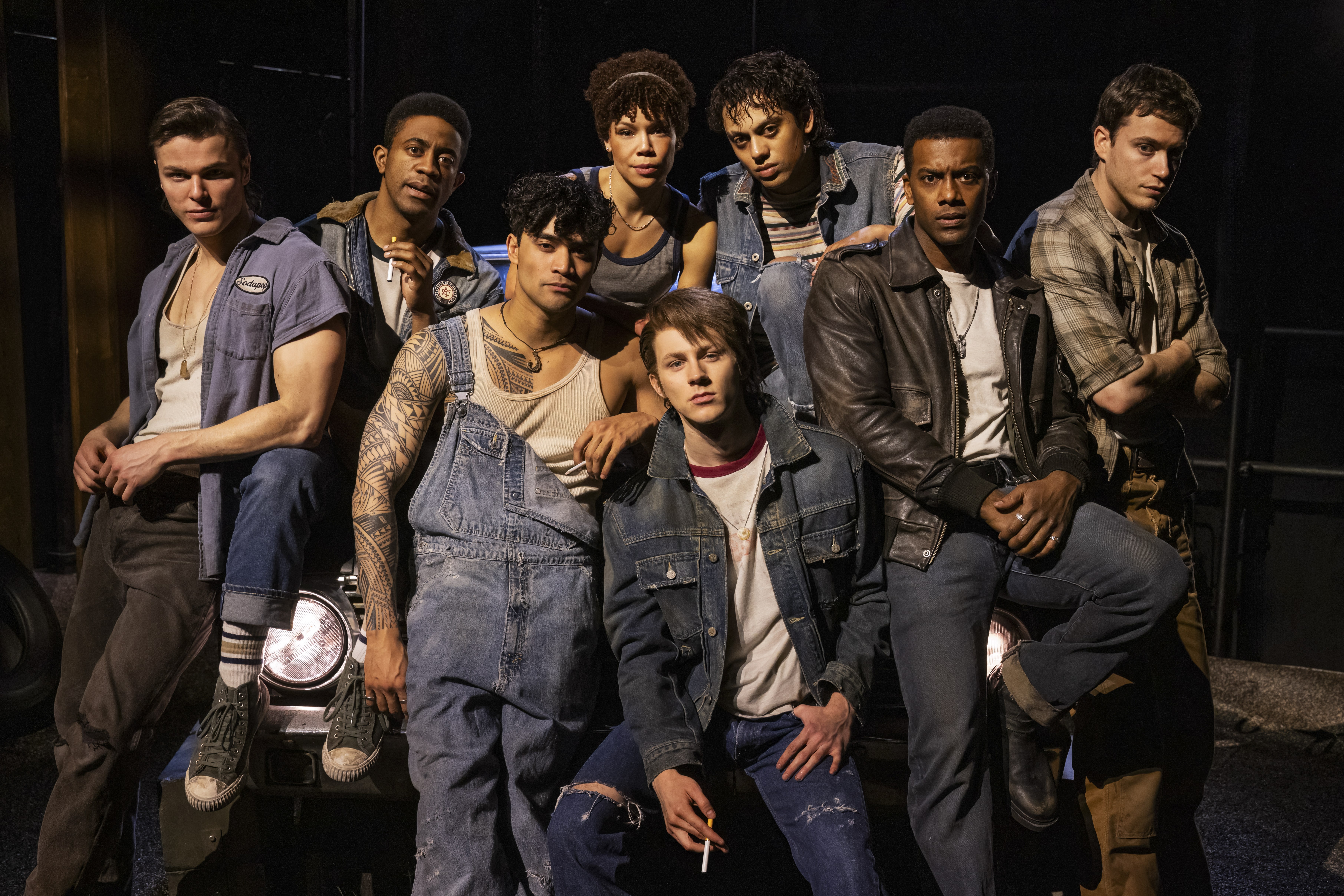 Promotional group portrait of seven stage performers posed closely together in casual denim, leather, and workwear-inspired costumes. The ensemble is arranged on and around a vehicle under dramatic theater lighting against a dark stage background.