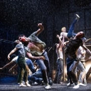 Production photo from The Outsiders showing three performers standing under a dramatic spotlight on a dark stage. Wearing casual, mid-century-inspired clothing including jeans, T-shirts, and a denim vest, the performers walk forward across a brightly lit section of the set while the surrounding stage remains in shadow.