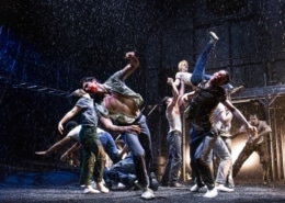 Production photo from The Outsiders showing three performers standing under a dramatic spotlight on a dark stage. Wearing casual, mid-century-inspired clothing including jeans, T-shirts, and a denim vest, the performers walk forward across a brightly lit section of the set while the surrounding stage remains in shadow.