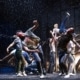 Production photo from The Outsiders showing three performers standing under a dramatic spotlight on a dark stage. Wearing casual, mid-century-inspired clothing including jeans, T-shirts, and a denim vest, the performers walk forward across a brightly lit section of the set while the surrounding stage remains in shadow.