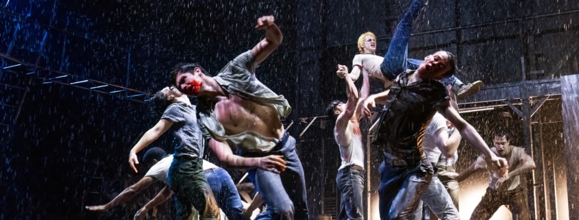 Production photo from The Outsiders showing three performers standing under a dramatic spotlight on a dark stage. Wearing casual, mid-century-inspired clothing including jeans, T-shirts, and a denim vest, the performers walk forward across a brightly lit section of the set while the surrounding stage remains in shadow.
