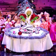 A green furry creature dressed in a santa suit carves into an illustrated turkey on top of a large, round table. Other colorful actors in pink costumes surround the table