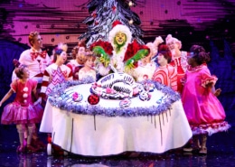 A green furry creature dressed in a santa suit carves into an illustrated turkey on top of a large, round table. Other colorful actors in pink costumes surround the table